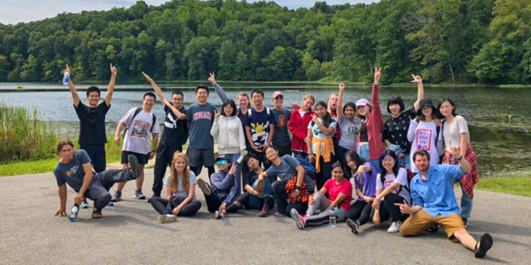 A large group of students pose for a goofy group portrait in front of a lake and wooded hills.