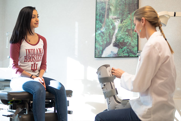 A female student sits on an examination table facing a female doctor holding a protective ankle and foot boot.